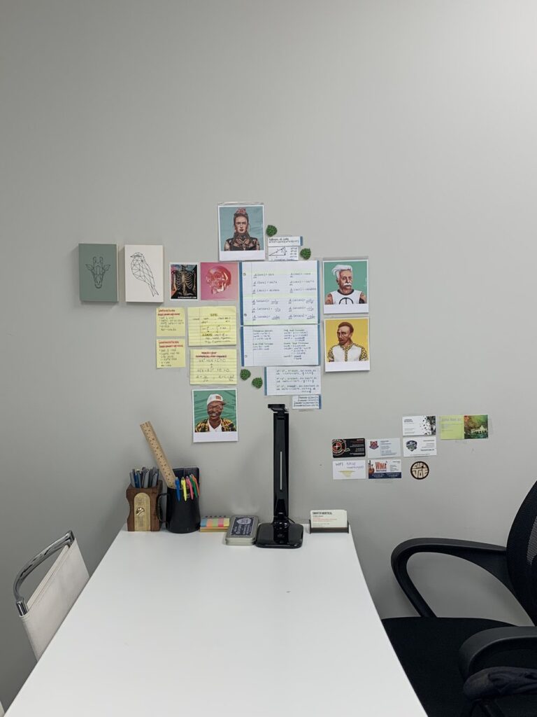 a long, white table with school supplies on it, one chair on each side facing each other, behind it are math equations on stick notes against the wall, with geometric art, and polaroid-style images of famous Albert Einstein, Vincent Van Gogh, Martin Luther king, and Frida Kahlo